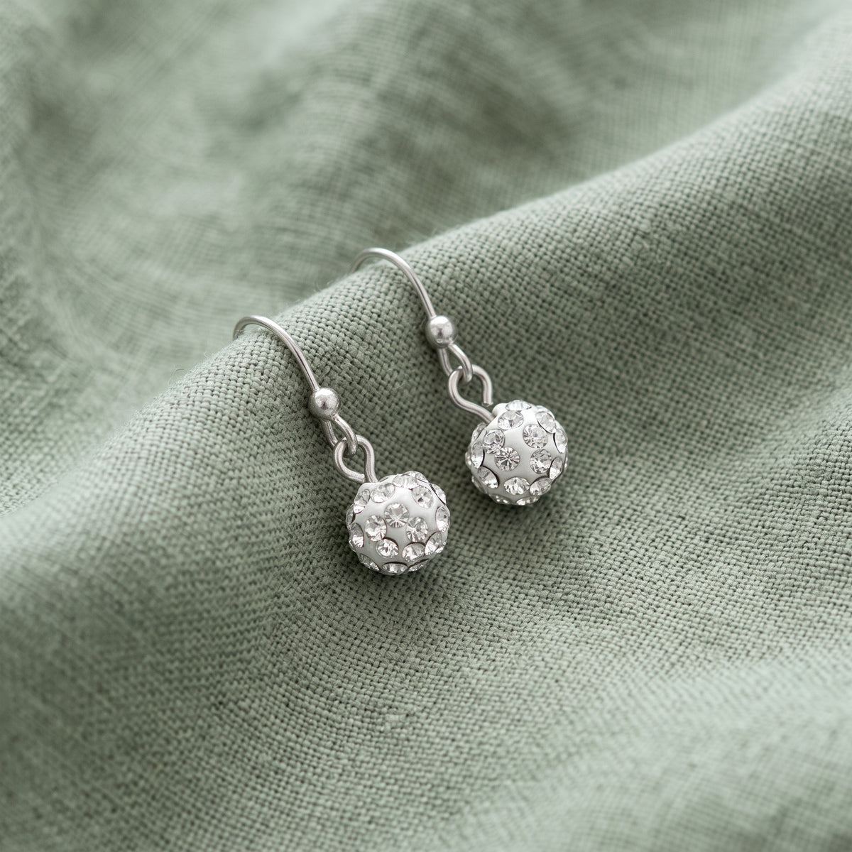 Crystal Fireball Pave 6mm Drop Earrings resting on sage green linen