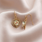 Gold earrings with heart design and porcelain rose details on a soft pink fabric background
