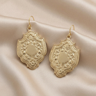 Ornate gold medieval shield earrings on a beige fabric background