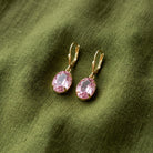 A pair of gold lever back earrings with oval pink crystals, displayed on a textured olive green fabric background.