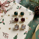 Three pairs of gold-toned stud earrings with green, red, and clear crystals, arranged on a Christmas-themed gift box with holly and pine decorations.