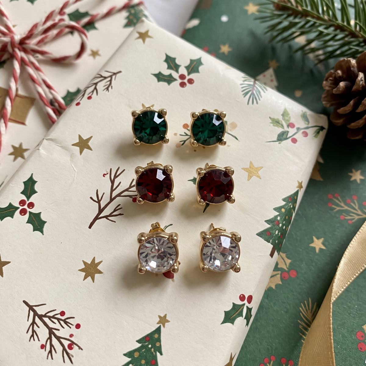 Three pairs of gold-toned stud earrings with green, red, and clear crystals, arranged on a Christmas-themed gift box with holly and pine decorations.