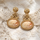 Antiquities Couture Carnelian Color Cameo Twin Muse And Bird Dangle Earrings, displayed on white lace fabric.