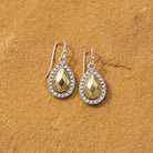 A pair of teardrop-shaped drop earrings with faceted gold tone centers and silver settings, displayed on a textured brown surface.