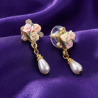 Decorative earrings with porcelain rose designs and faux pearl drops on a purple fabric background