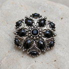 Jet Black Stone Ornate Brooch Pin displayed on a textured light beige stone surface.