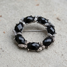 Black Faceted Stone Wreath Pin, photographed on a textured light gray surface.