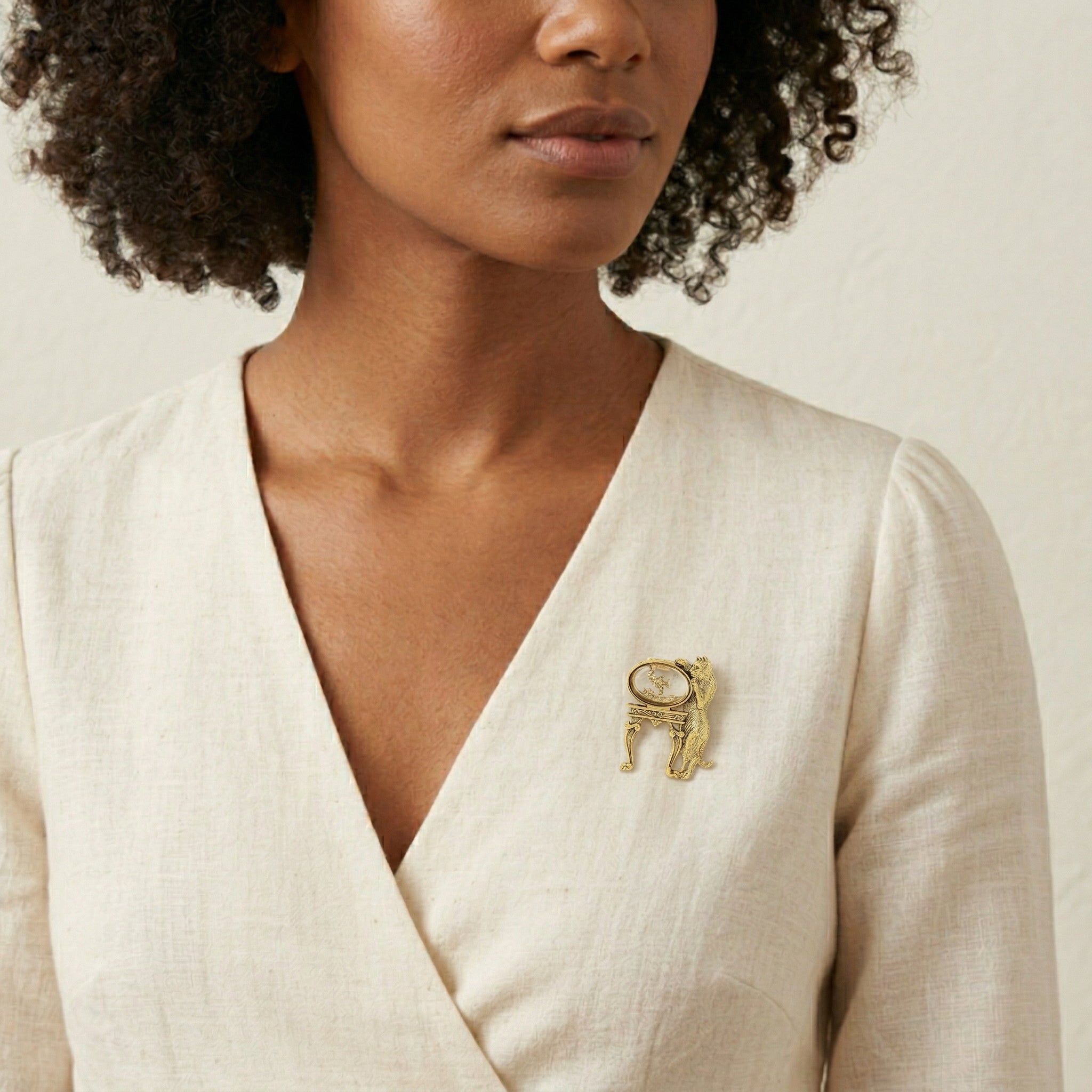 Woman wearing a gold tone fish bowl and cat brooch on a beige background