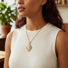 Woman wearing a gold necklace with a floral pendant in a home setting.