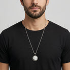 Man wearing a black t-shirt with a silver necklace featuring a round white stone pendant.