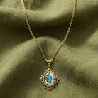 A gold chain necklace with an oval pendant featuring a wine grapevine decal, displayed on draped olive green fabric.
