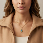 A close-up of a woman wearing a gold wine grape decal pendant necklace with a blue  iridescent stone, styled with a camel-colored zip-up jacket.