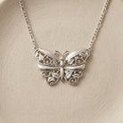A silver butterfly pendant necklace with intricate filigree detailing, displayed on a textured beige surface.