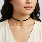 Woman wearing a black choker with a gold pendant that has two charms on a plain background
