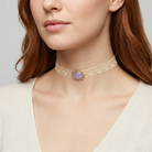 Close-up of a woman wearing a gold choker necklace with a purple gemstone on a neutral background