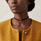 Person wearing a black choker with a gold clasp, wearing a mustard yellow textured garment.