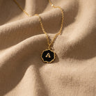Sunburst Black Enamel Initial Pendant Necklace resting on beige linen fabric
