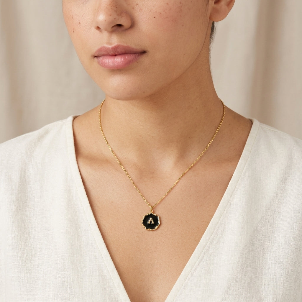 Gold tone necklace with a black lettered initial pendant worn by a person in a white top against a neutral background