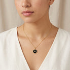Gold tone necklace with a black lettered initial pendant worn by a person in a white top against a neutral background