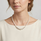 A close-up of a woman wearing a pearl necklace with a silver-toned chain and a row of larger pearls at the front, paired with a cream-colored top against a light gray background.