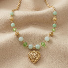 A gold filigree heart pendant necklace with light green and milky aventurine beads, displayed on a soft beige fabric background.