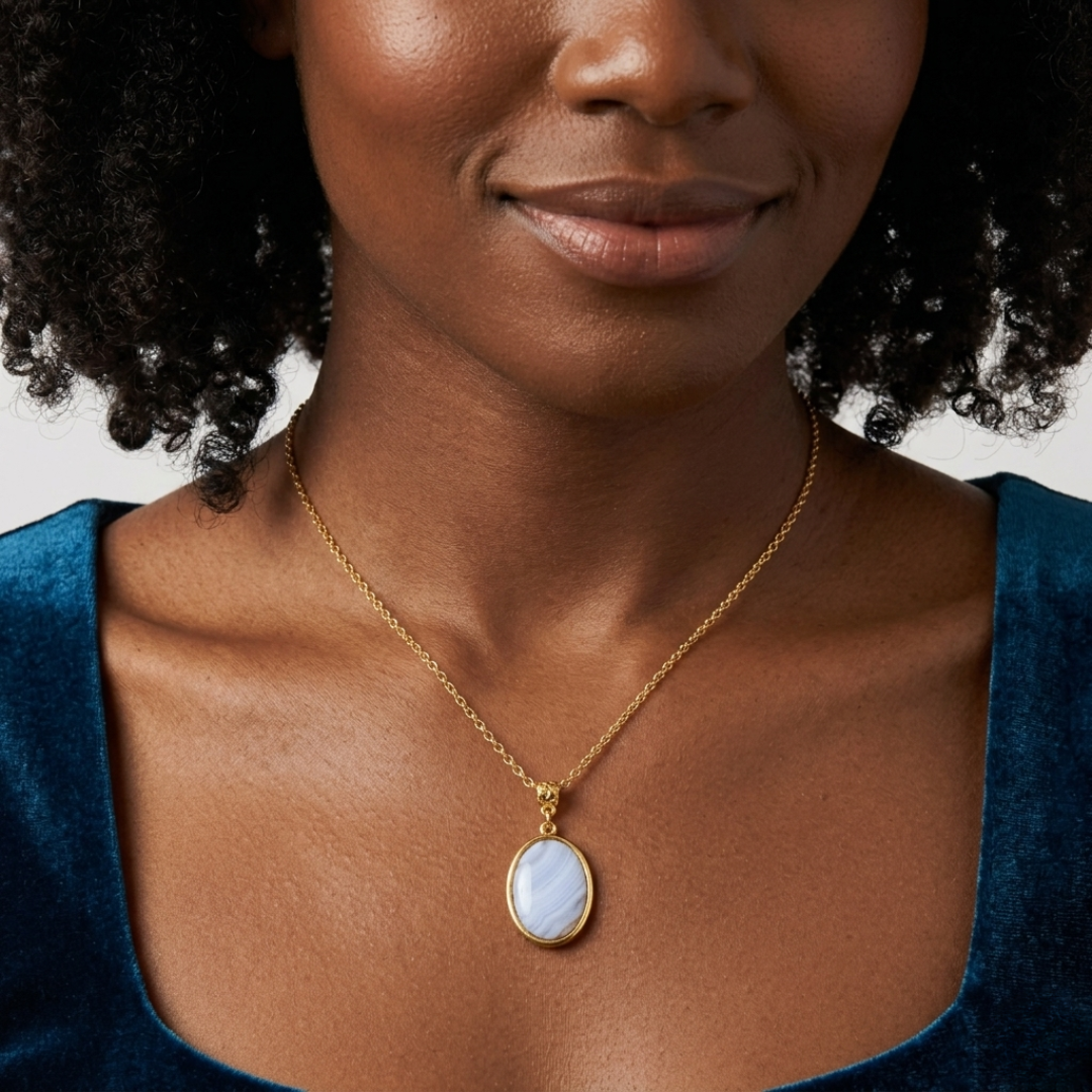 Woman wearing a gold necklace with an oval blue agate pendant on a plain background