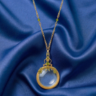 Gold necklace with a magnifying glass pendant and green crystal accents on a blue fabric background