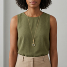 Woman wearing a green sleeveless top and beige pants with a gold magnifying glass necklace against a neutral background