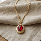 Gold tone locket necklace with a red stone pendant on beige fabric