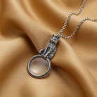 Silver cat magnifying glass pendant necklace on a chain against a beige fabric background