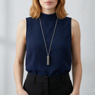 Person wearing a navy sleeveless top with a long vial pendant necklace against a plain background