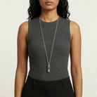 Woman wearing a gray sleeveless top and black pants with a long whistle pendant necklace on a plain background