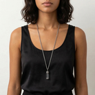Woman wearing a black top and a long necklace with a glass vial pendant against a plain background