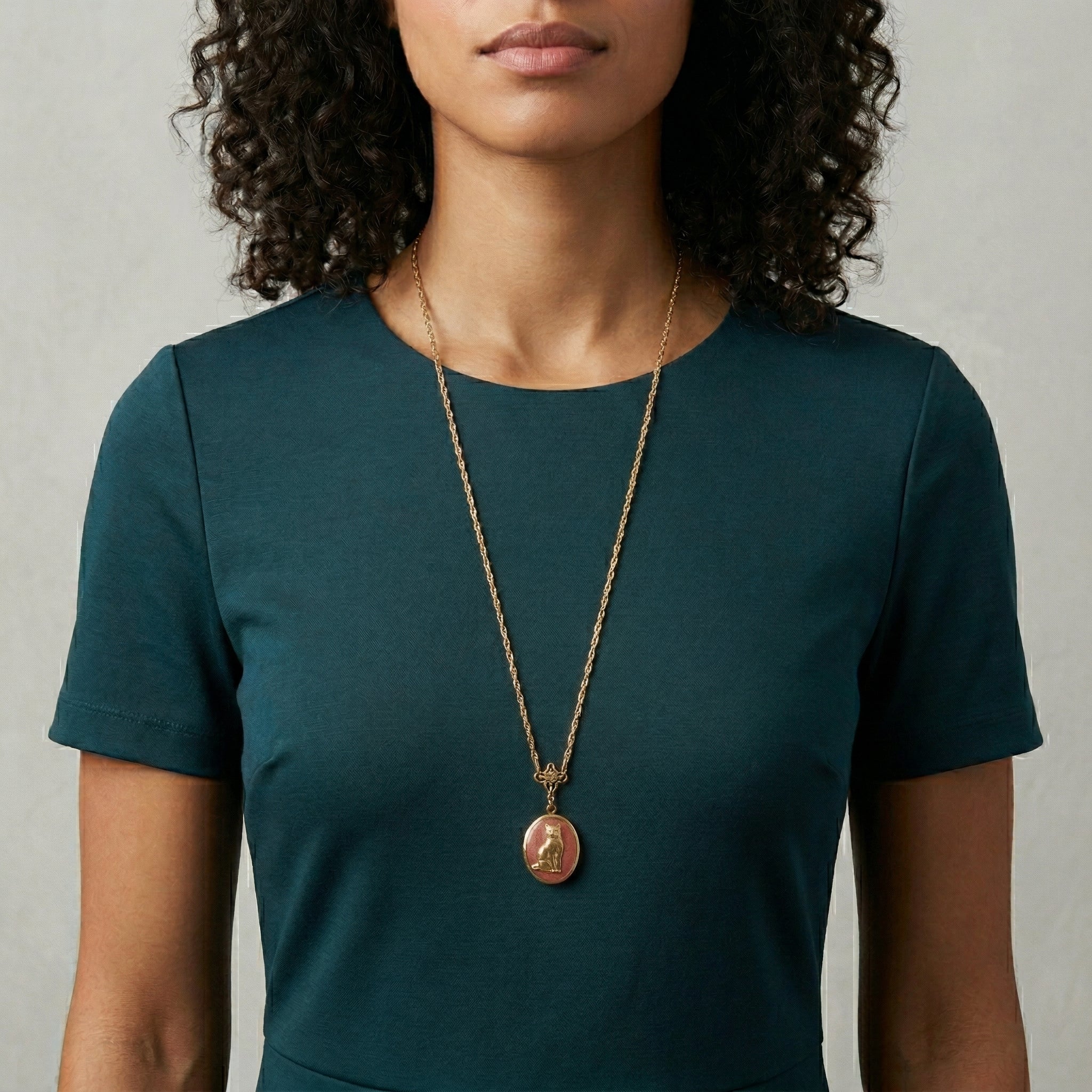Woman wearing a green shirt and a long necklace with a cat locket pendant against a neutral background