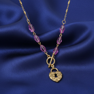 Gold necklace with purple beads and a heart-shaped lock pendant on a blue fabric background