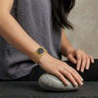 Person wearing a gold bracelet with a dark stone on a gray background