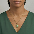 Woman wearing a gold necklace with a green pendant against a neutral background