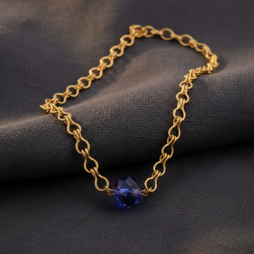 Gold chain bracelet with a blue bead pendant on a dark fabric background