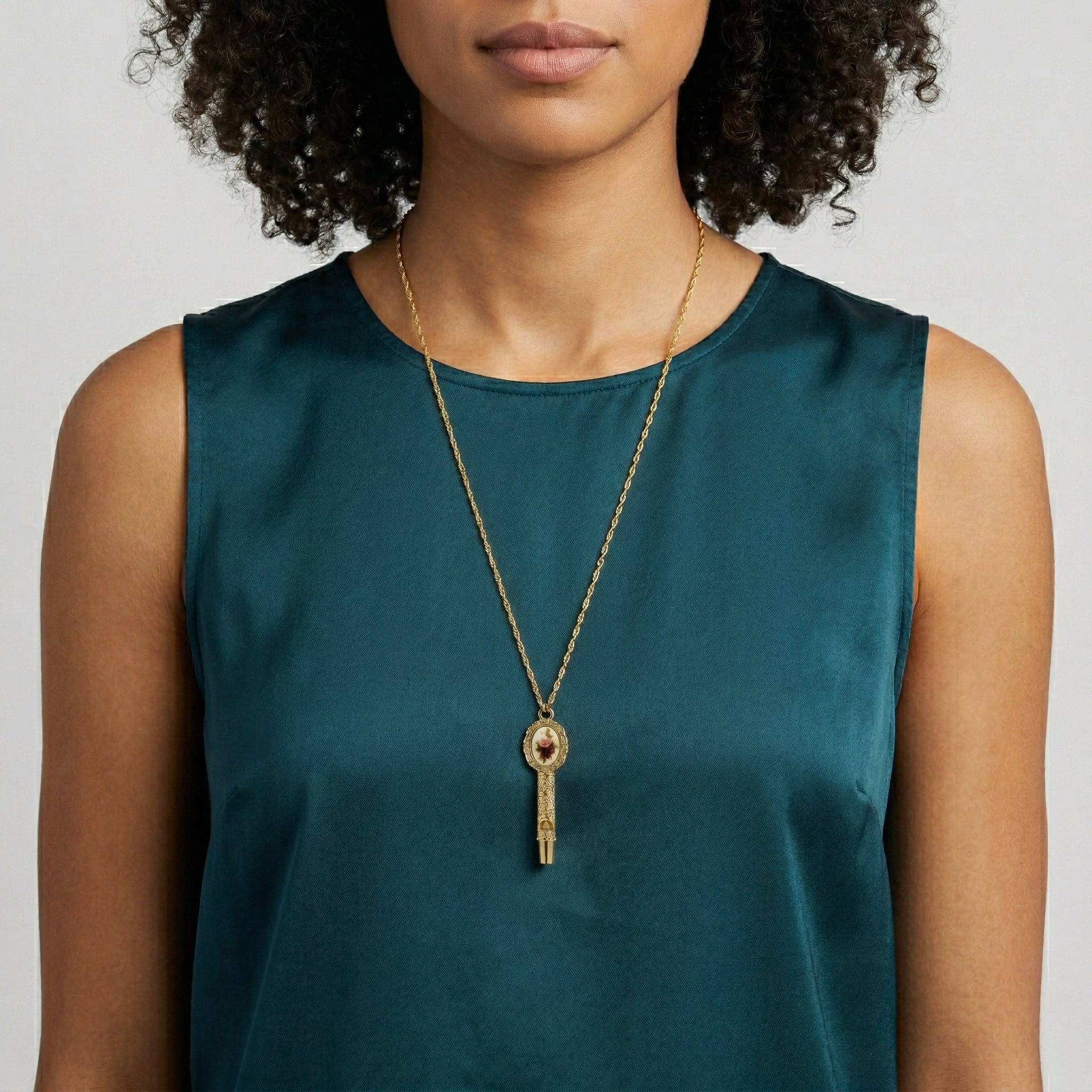 Woman wearing a green sleeveless top and a gold necklace with a floral whistle pendant on a neutral background