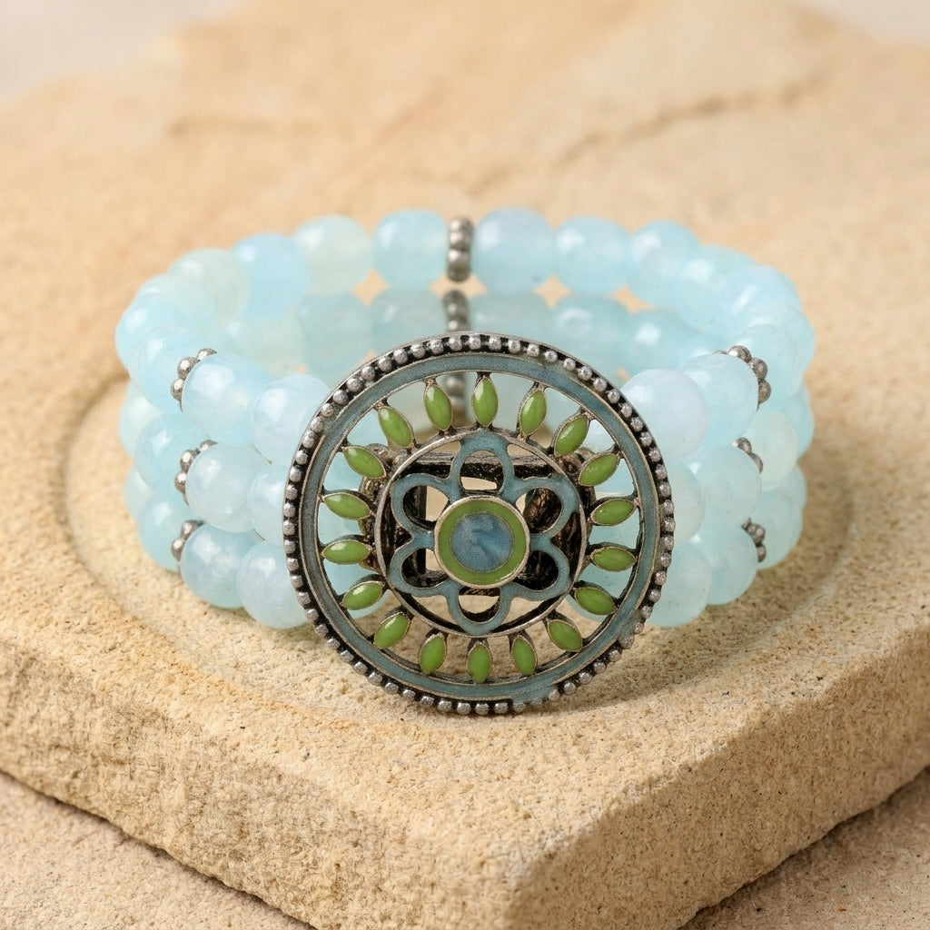 Light blue beaded bracelet with a decorative green and silver circular clasp on a stone surface