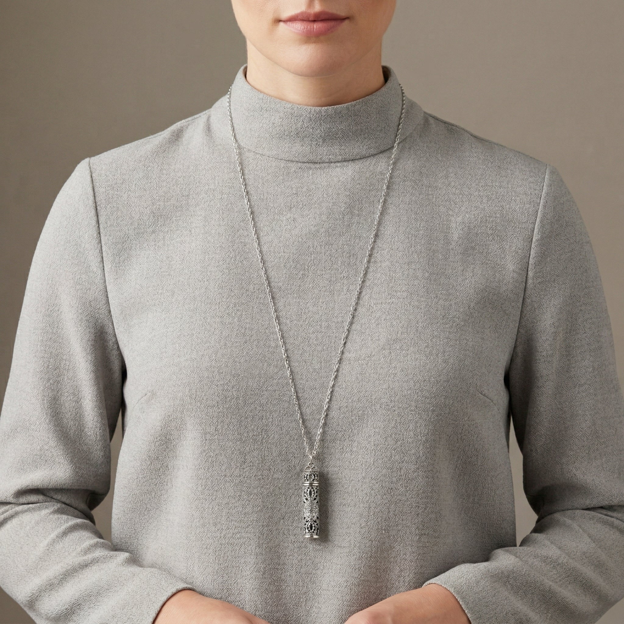 Person wearing a gray sweater with a long necklace with a vial pendant against a neutral background
