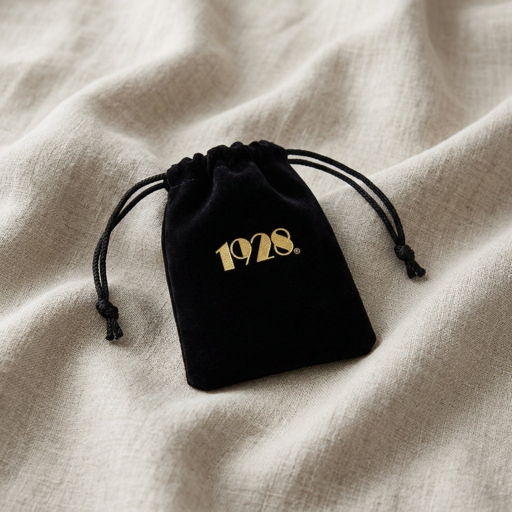 Black velvet drawstring jewelry pouch with golden 1928 logo resting on linen cloth surface
