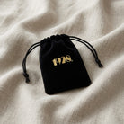 Black velvet drawstring jewelry pouch with golden 1928 logo resting on linen cloth surface