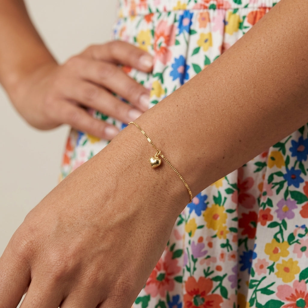 A delicate gold chain bracelet with a small puffed heart charm worn on a person's wrist, shown against a colorful floral dress and neutral background.