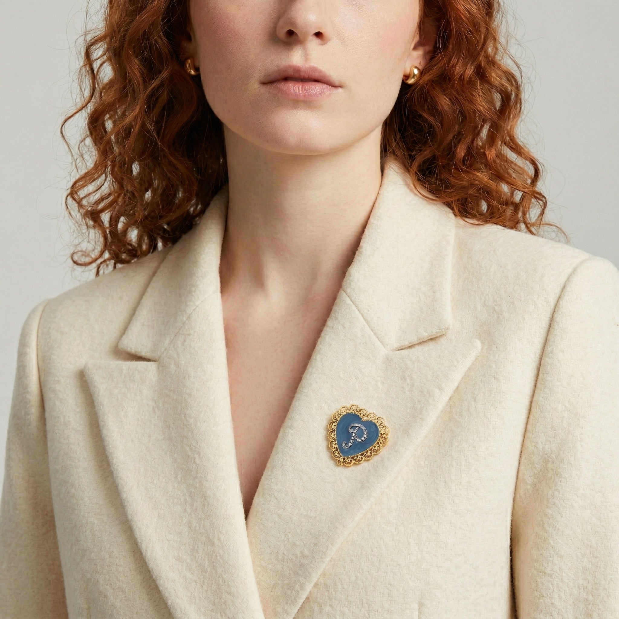 Woman wearing a cream blazer with a decorative initialed brooch on a neutral background