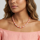 A close-up of a woman wearing a Peach Cracked Glass & White Faux Pearl Necklace 15" + 3" Extension, paired with a matching off-the-shoulder top against a light gray background.