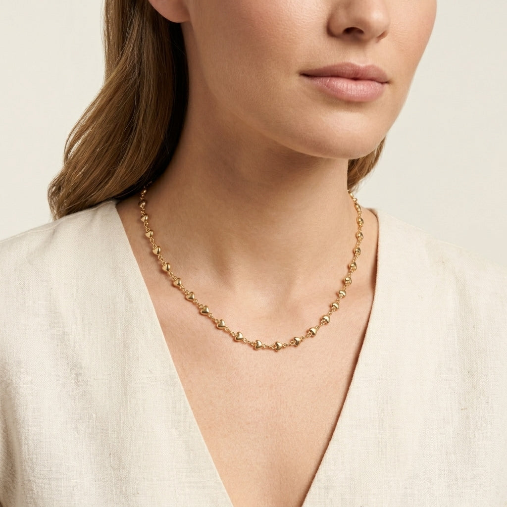 A close-up of a woman wearing a delicate gold beaded necklace with smaller heart shaped beads, paired with a light beige linen top against a soft cream background.