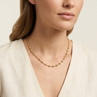 A close-up of a woman wearing a delicate gold beaded necklace with smaller heart shaped beads, paired with a light beige linen top against a soft cream background.