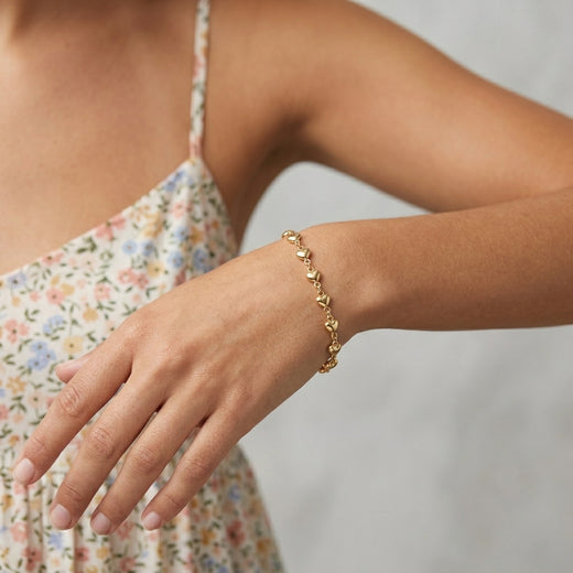 A delicate Classic Golden Heart Link Chain Bracelet worn on a woman's wrist. The model is wearing a light floral print camisole against a soft, light gray background.