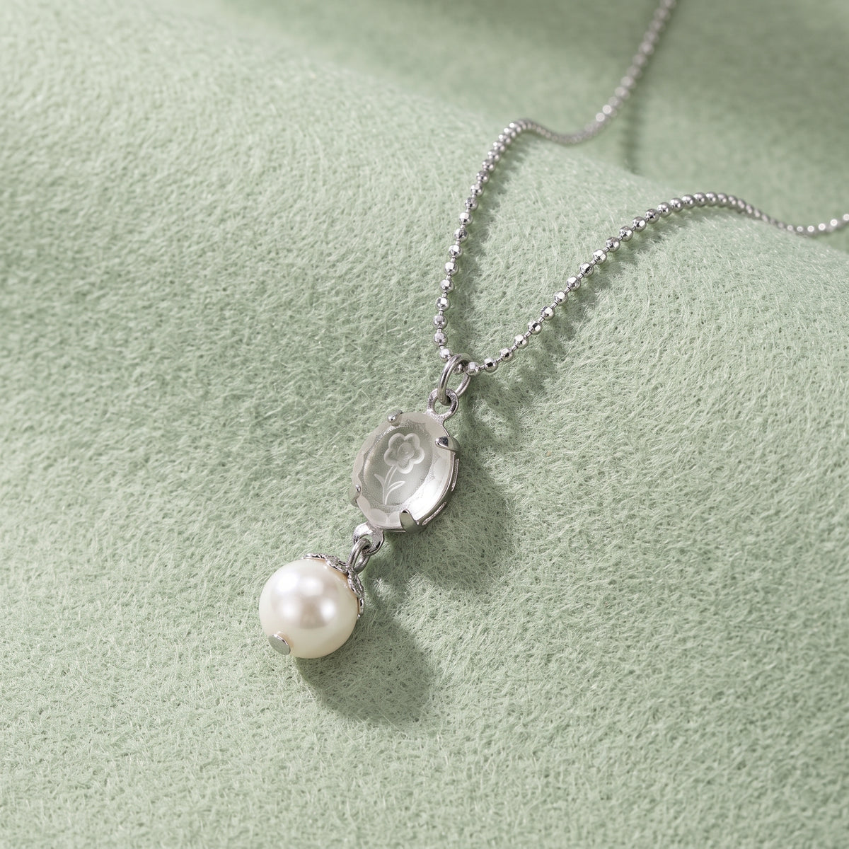 Translucent Flower Engraved Stone & Faux Pearl Drop Pendant Necklace displayed on a soft light green textured fabric background.